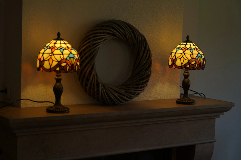Deux lampes de table Tiffany allumées, en verre jaune, orange et turquoise, disposées symétriquement sur une cheminée en pierre avec une grande décoration murale ronde en osier entre elles.