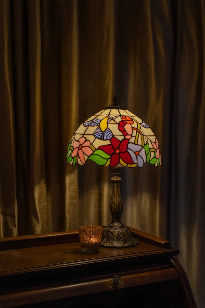 Lampe de table Tiffany aux tons chauds ambrés et verts sur une commode, projetant une douce lueur colorée sur le mur et apportant chaleur et nostalgie à la maison.