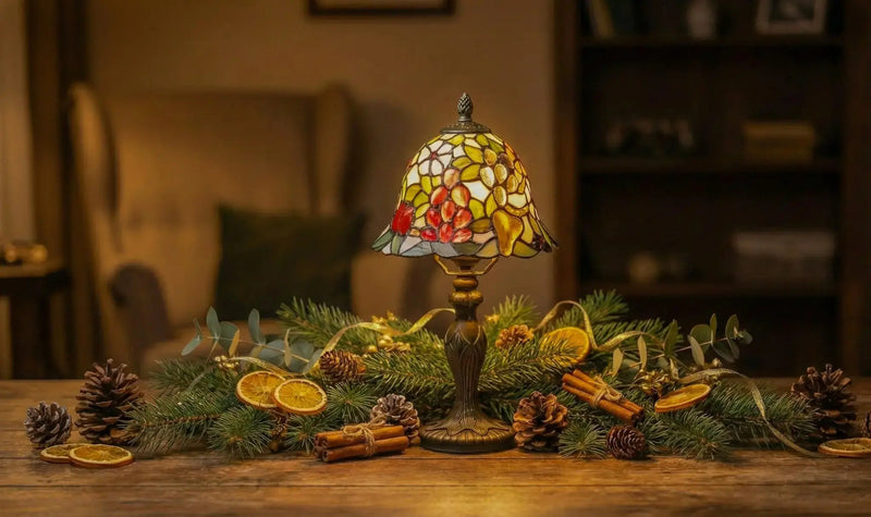 Lampe de table Tiffany avec abat-jour en vitrail coloré sur une table en bois, entourée de branches de sapin, pommes de pin, tranches d'orange séchées et cannelle, dans un salon chaleureux.
