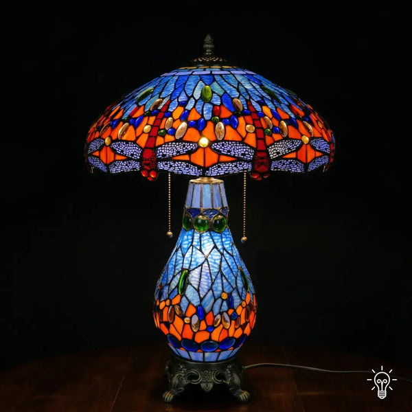 Lampe de table Tiffany allumée, abat-jour en dôme avec zone supérieure en vitrail bleu, zone centrale orange-rouge avec cabochons en verre bleus et ambrés, bordure de libellules bleu lavande en bas ; pied bleu clair en forme de poire et base en bronze à pattes de lion