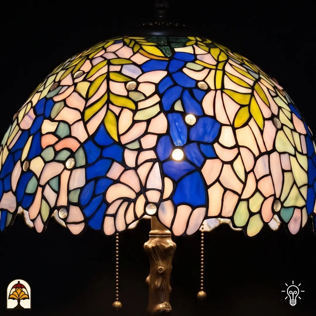 Gros plan abat-jour allumé lampe de table Tiffany Wisteria Celeste – grappes de glycine bleues et feuilles vert-jaune en vitrail, pied en bronze en forme de tronc d'arbre