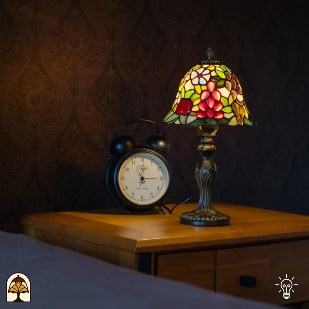 Lifestyle — lampe de table Tiffany allumée sur une table de chevet en bois à côté d'un réveil vintage, papier peint sombre à petites fleurs en arrière-plan