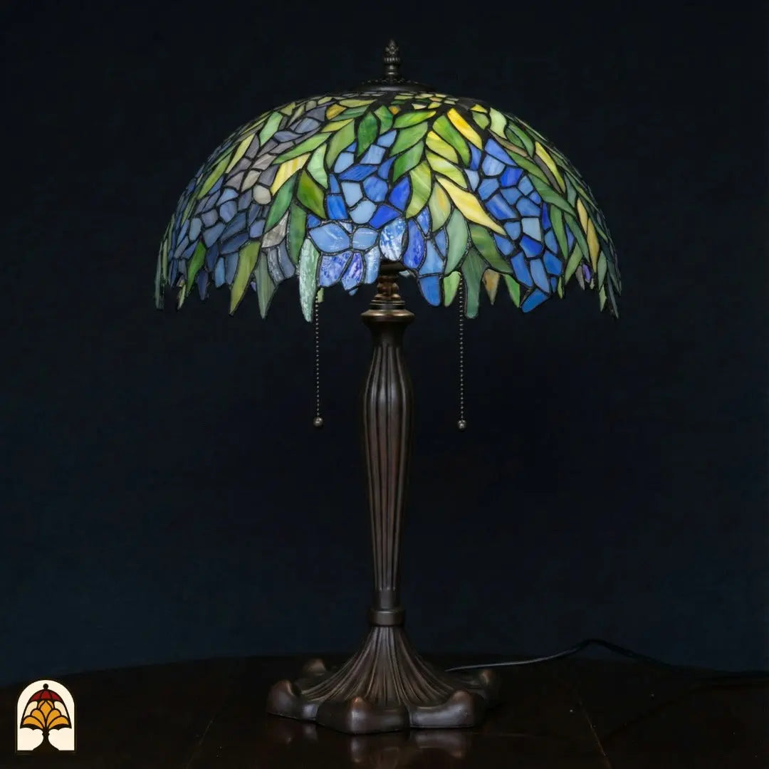 Lampe de table Tiffany éteinte sur fond bleu foncé — large abat-jour en dôme avec feuilles vertes et fleurs bleues, pied en bronze avec interrupteurs à chaînette