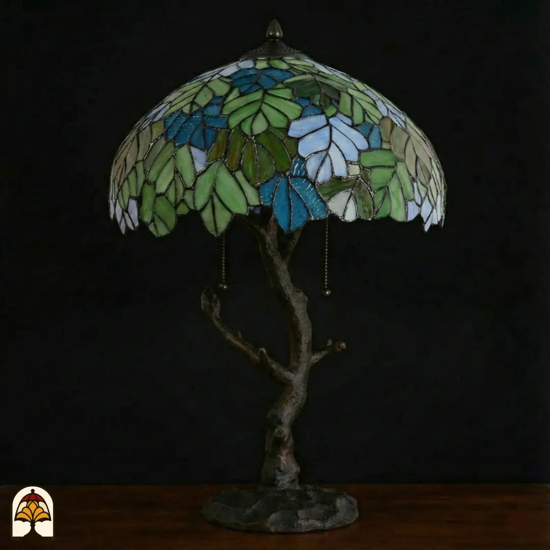 Lampe de table Tiffany Sylva Verde éteinte sur fond sombre, abat-jour dôme à motifs de feuilles vert-bleu et blanches en vitrail, pied en forme de tronc d'arbre avec interrupteurs à chaînette
