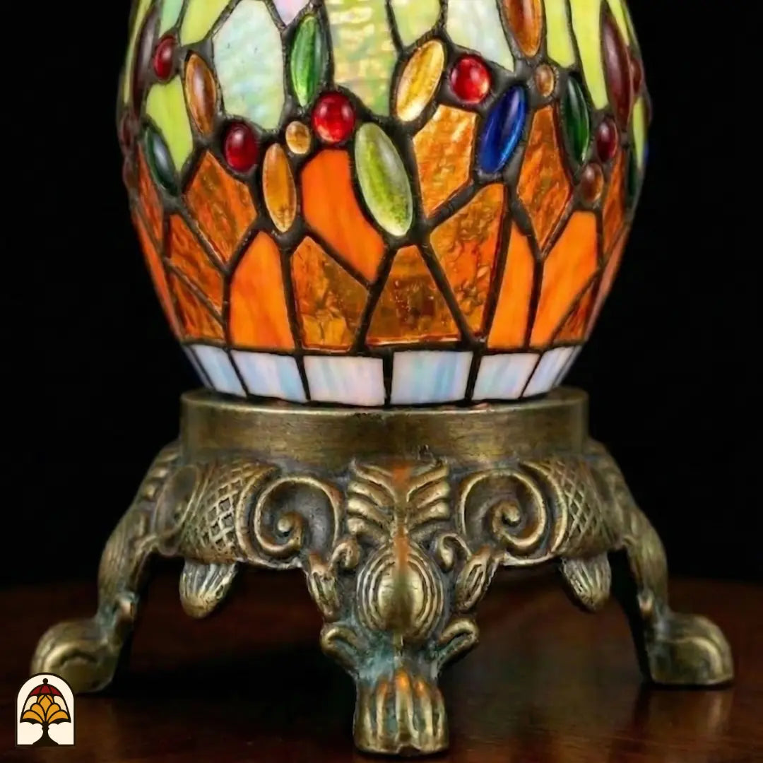 Gros plan pied décoratif en bronze Libellula Lumina ⌀30 avec pied sphérique en vitrail, panneaux orange et verts avec cabochons en verre colorés, socle en bronze à motifs de feuilles d'acanthe et de volutes et pattes de lion