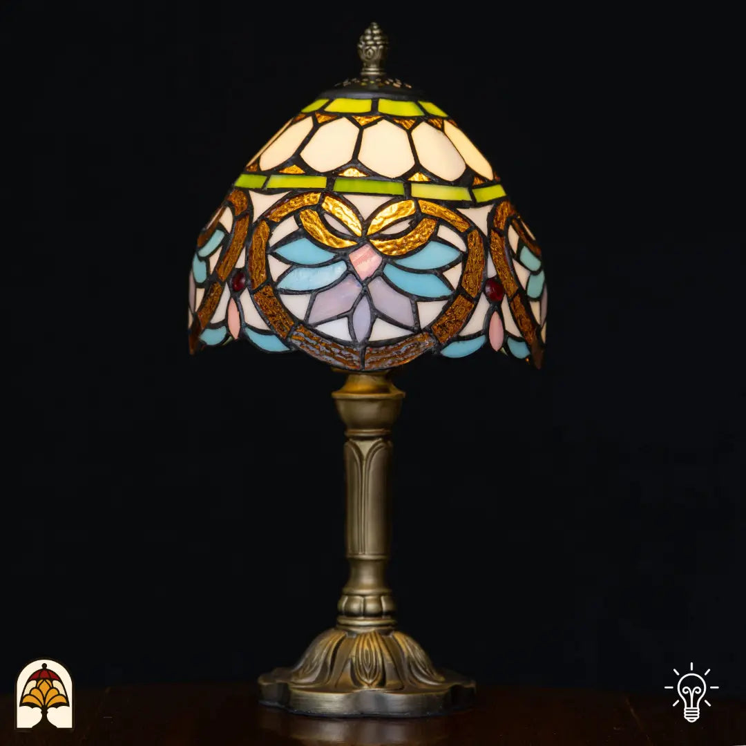 Lampe de table Tiffany Lucette ⌀20 cm allumée sur fond noir – abat-jour en dôme crème avec bandeau décoratif vert, formes dorées de libellules et de feuilles, pétales bleu clair et roses, pierres précieuses rouges, pied colonne en bronze