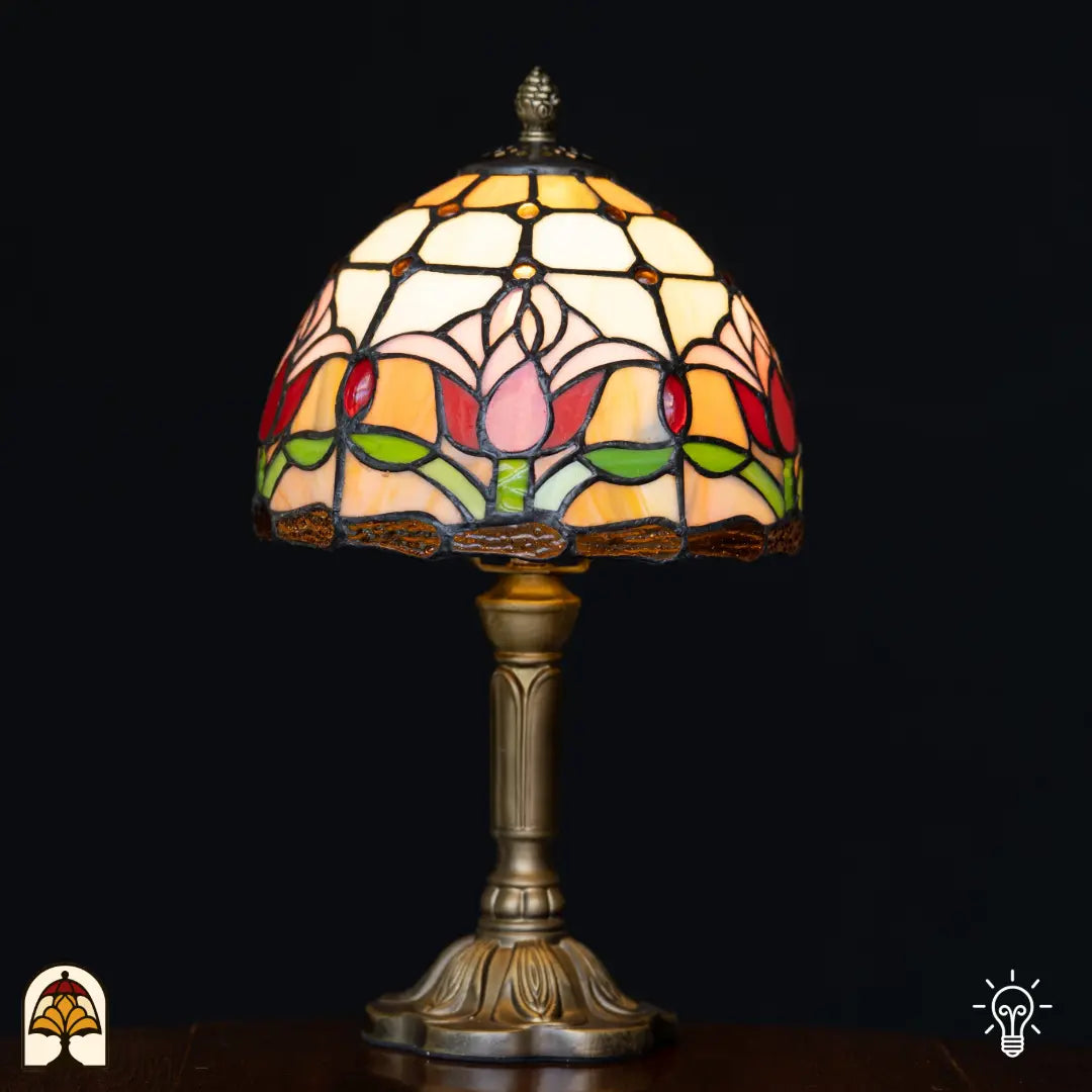 Lampe de table Tiffany Tulipa ⌀20 cm allumée sur fond noir – abat-jour en dôme avec tulipes roses et rouges sur vitrail crème et orange, pied colonne en bronze