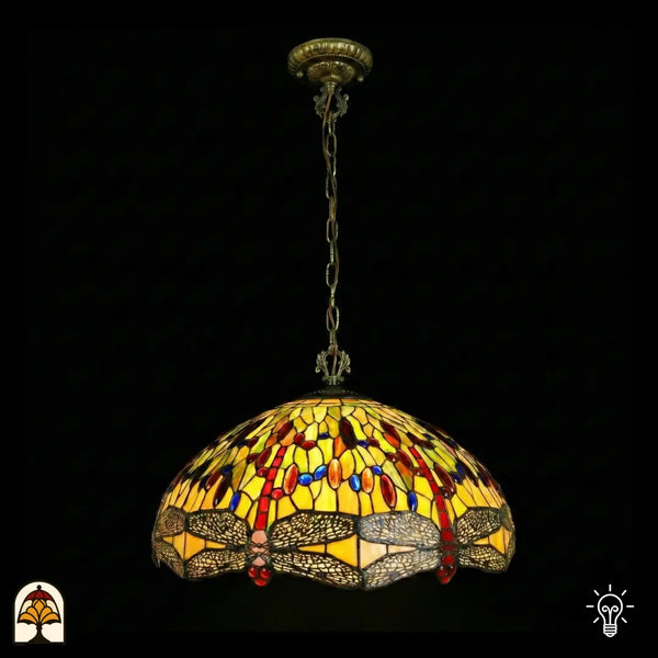 Suspension Tiffany Libellula Aurelia ⌀50 éteinte sur fond sombre, grand abat-jour en dôme à panneaux de vitrail jaunes, frise de libellules en bas, cabochons en verre rouges et bleus, chaîne en bronze et rosace de plafond