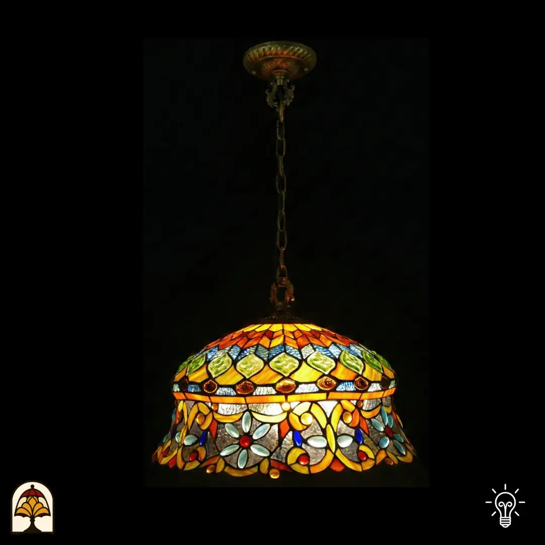 Suspension Tiffany allumée, abat-jour en dôme avec zone supérieure ambrée à cabochons en verre verts en losange et verre mosaïque bleu, zone inférieure avec arabesques jaunes et cabochons en forme de fleur bleu clair sur verre transparent, bord festonné, suspendue à une chaîne en bronze