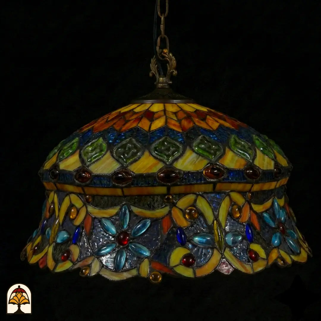Suspension Tiffany éteinte en gros plan, abat-jour en dôme avec zone supérieure ambre-jaune à cabochons en verre verts, et zone inférieure bleue à cabochons en forme de fleur bleu clair et cabochons en verre rouges sur verre gris-violet
