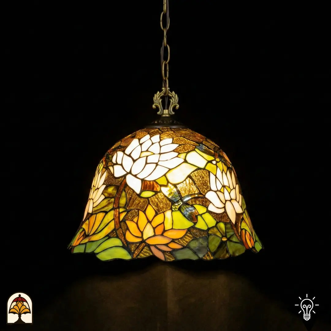 Suspension Tiffany allumée en gros plan, abat-jour en forme de cloche avec fleurs de lotus blanches en haut, fleurs de lotus orange-jaune en bas, feuilles de nénuphar vertes et libellule bleue sur fond ambre-brun, suspendue à une chaîne en bronze