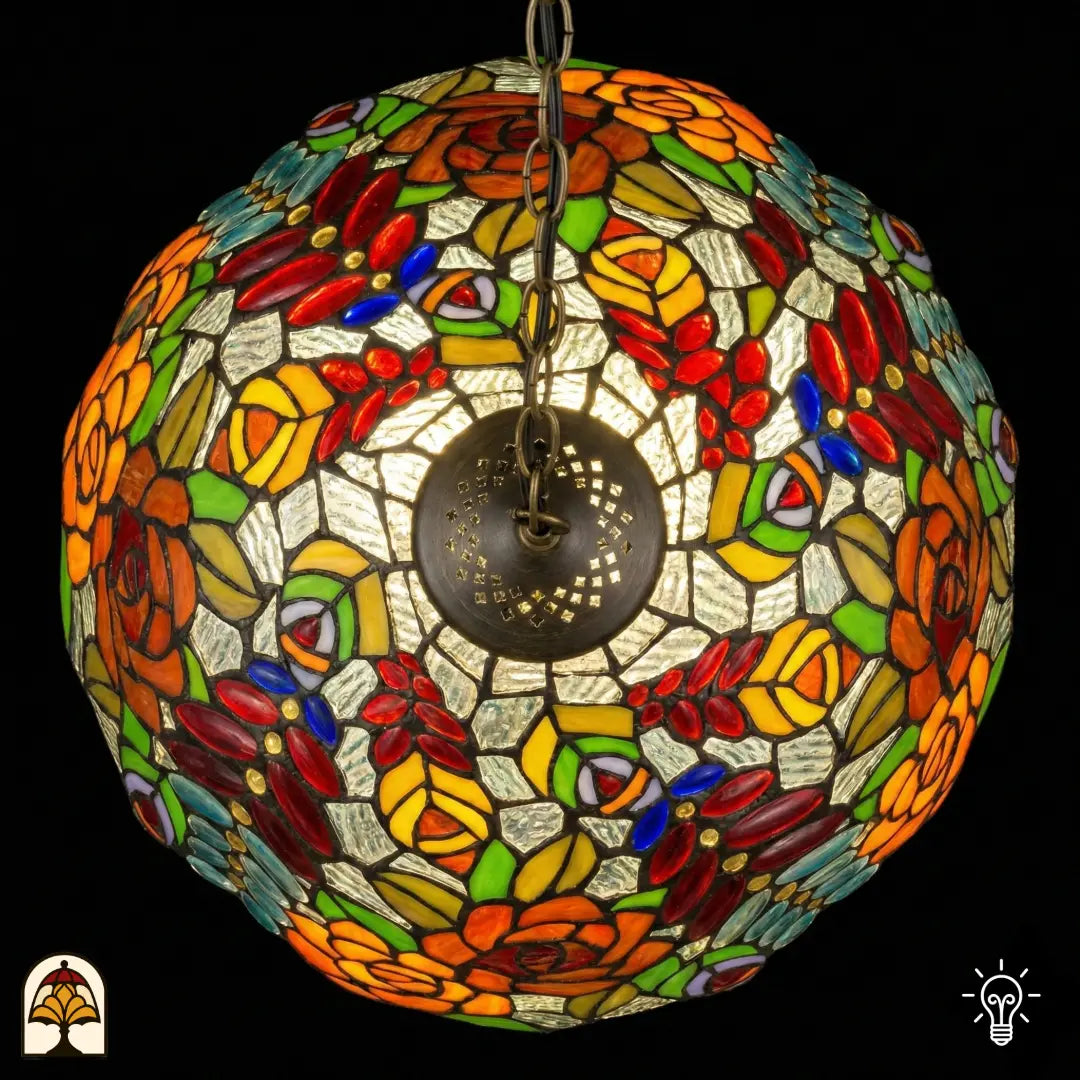 Vue de dessus de suspension lampe Tiffany allumée, dôme rond au motif symétrique de roses orange et jaunes, cabochons en verre rouges et feuilles vertes, plaque d'aération centrale en bronze visible