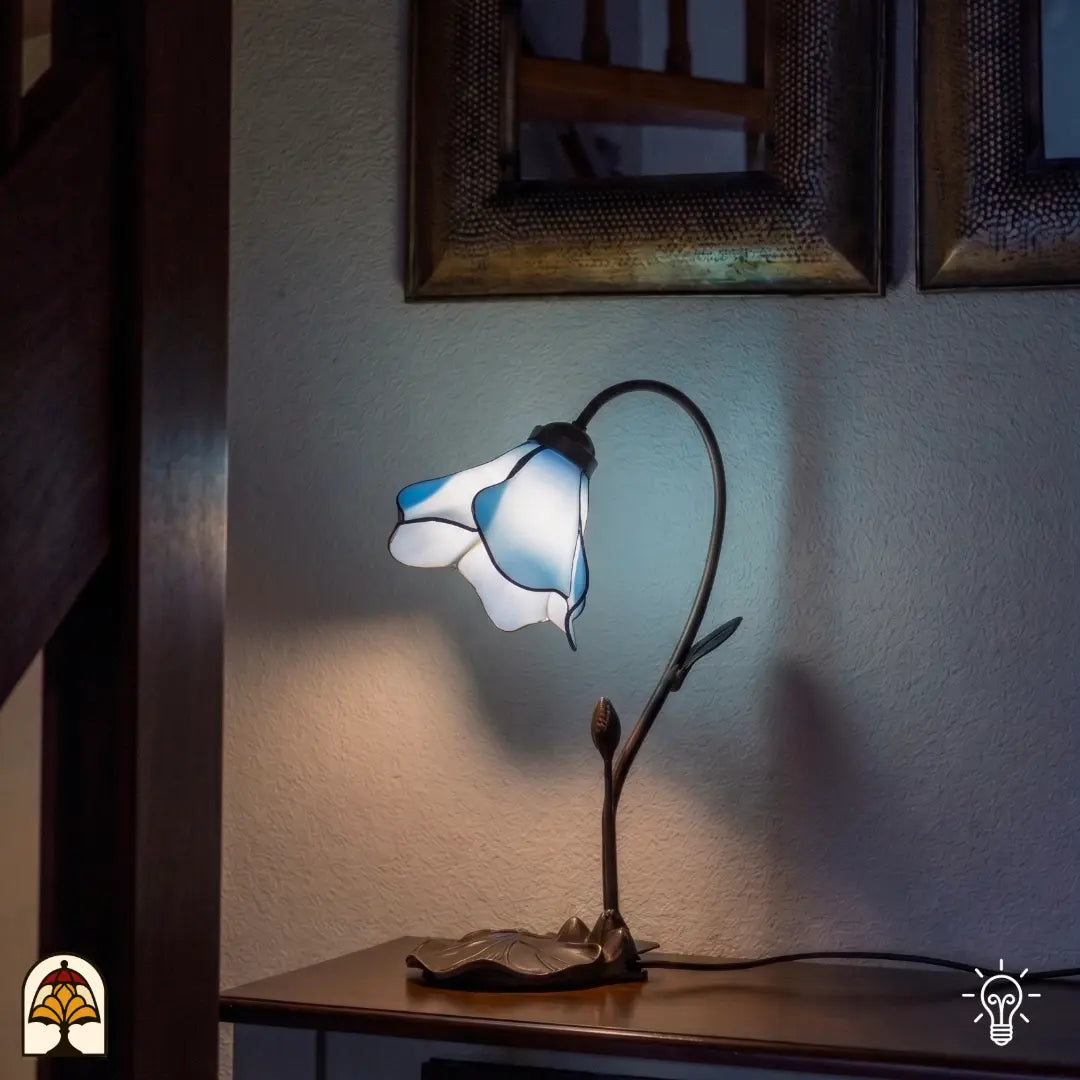 Une lampe de table Tiffany pleine d'atmosphère en forme de calice de fleur bleu et blanc, reposant sur un pied ouvragé couleur bronze à motifs de feuilles. La lampe est posée sur un meuble en bois contre un mur blanc, avec en arrière-plan deux miroirs décoratifs et un escalier en bois. La lampe diffuse une lumière douce et bleutée dans une pièce joliment éclairée.