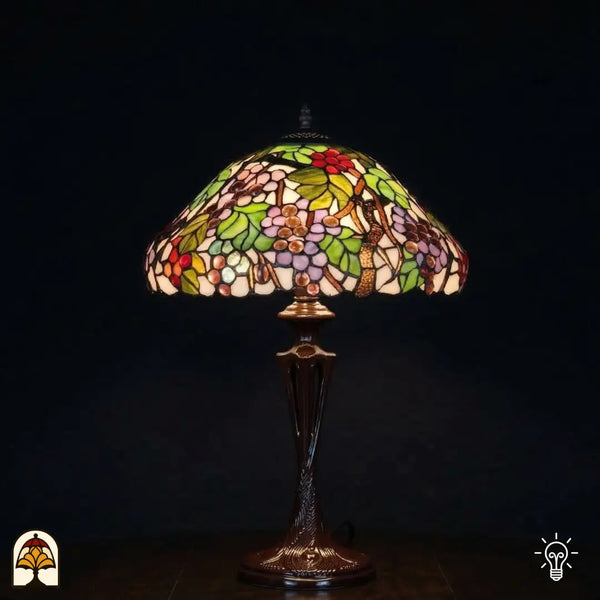 Lampe de table Tiffany Sylva Berria allumée avec large abat-jour en dôme, motifs de vigne avec petites grappes de raisins rouges et lilas et feuilles vertes en vitrail, pied vase Art nouveau en bronze avec interrupteurs à chaînette, fond sombre