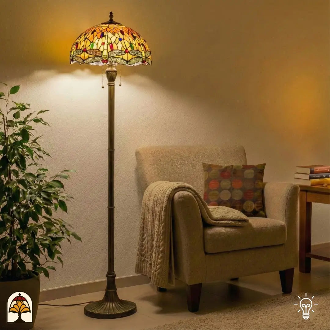 Lampadaire Tiffany Libellula Aurea allumé dans un salon — à côté d'un fauteuil beige avec plaid et coussin, plante verte à gauche, table d'appoint avec livres à droite