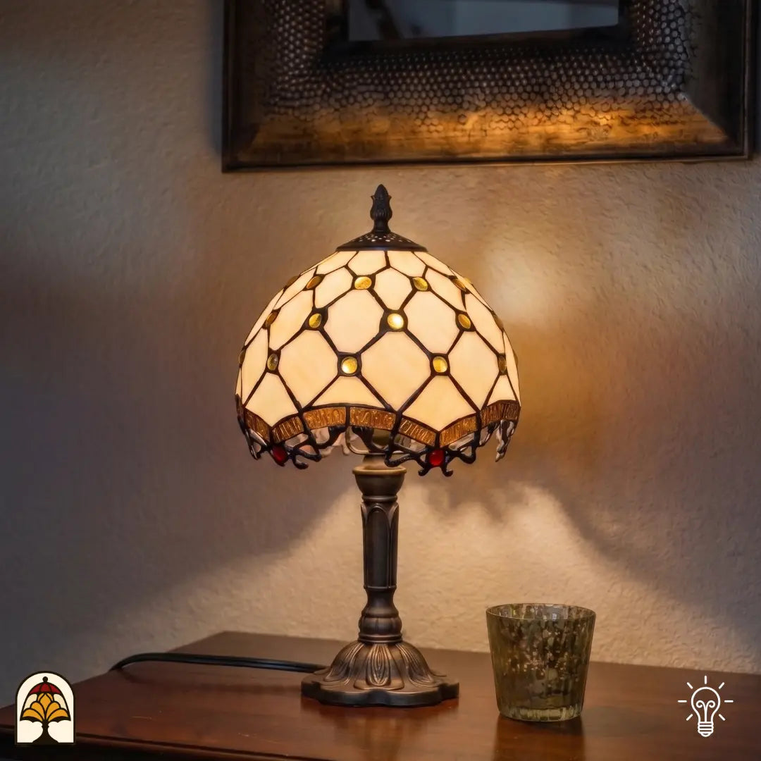 Lampe de table Tiffany Reticolo d'Oro ⌀20 cm allumée dans un intérieur chaleureux sur une table en bois – abat-jour crème avec cabochons en verre dorés et motif losange, pied en bronze, lumière chaude