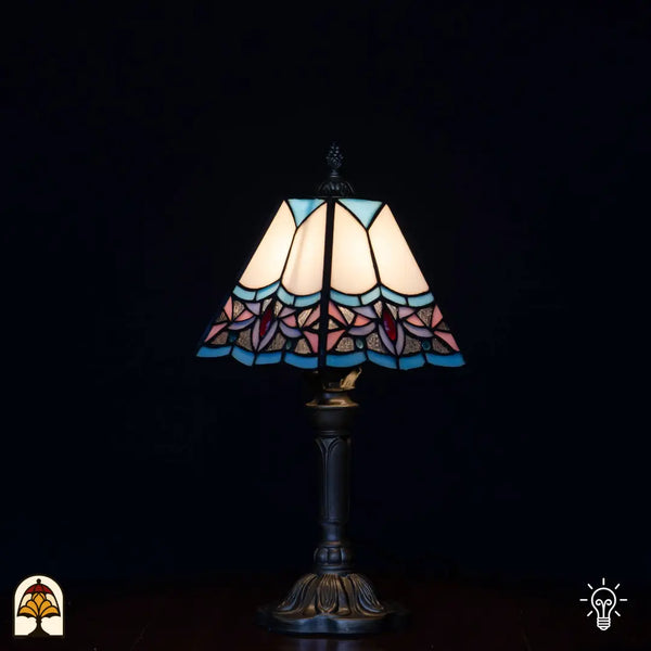 Lampe de table Tiffany allumée avec abat-jour trapézoïdal, panneaux supérieurs blancs/crème, bords bleu clair, fleurs de lotus roses et accents rouges en bas, pied chandelier classique, fond noir
