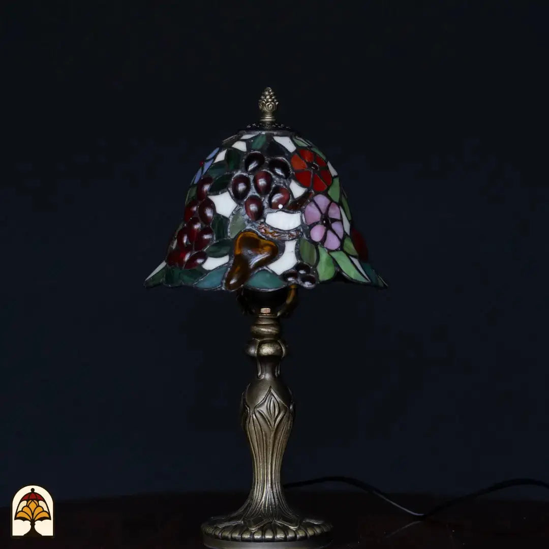 Lampe de table Tiffany éteinte sur fond sombre — abat-jour en forme de cloche avec raisins bordeaux, verre ambré, fleurs roses et bleues, feuilles vertes, pied chandelier classique