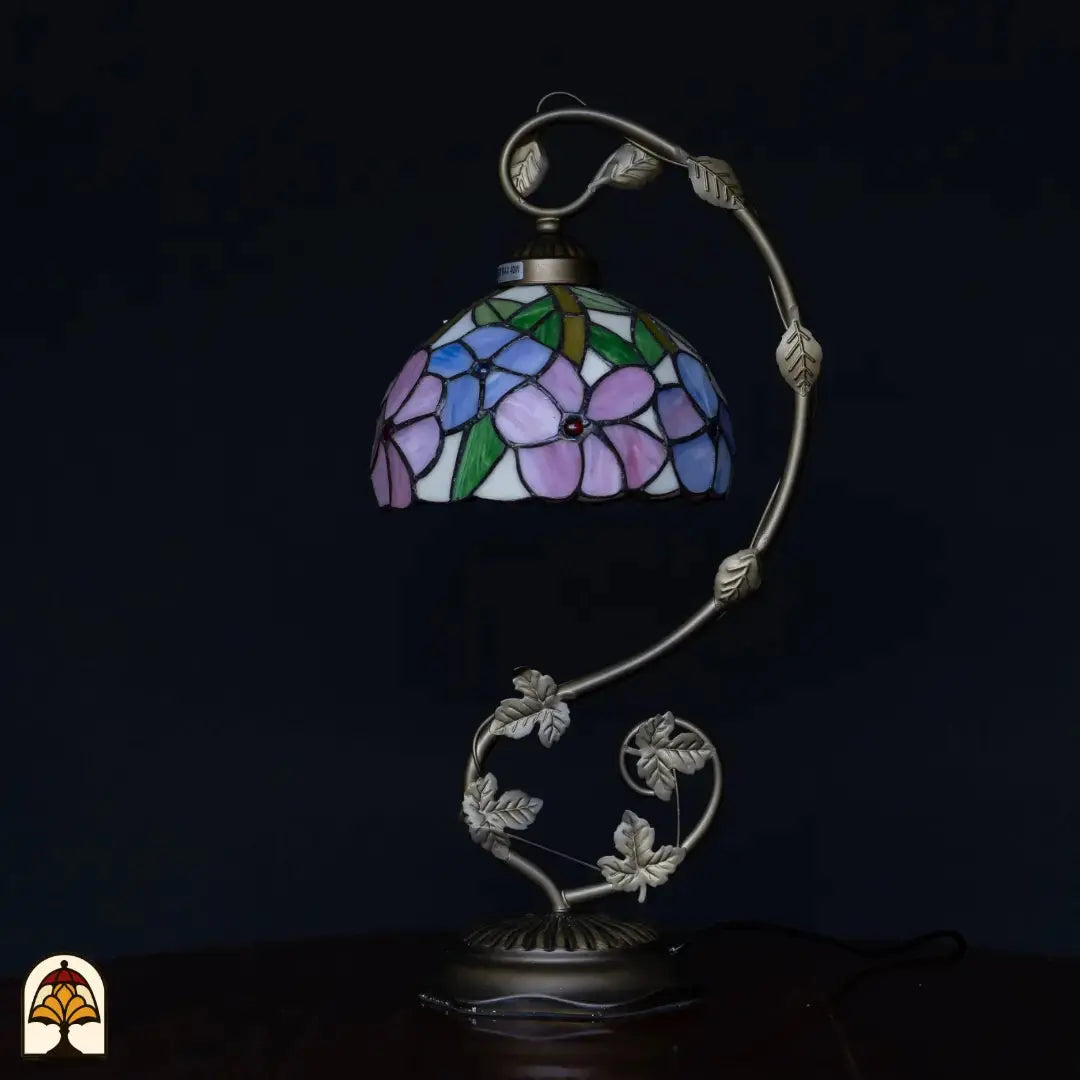 Lampe de table Tiffany éteinte sur fond sombre — abat-jour avec fleurs bleu/violet et roses, feuilles vertes, bras courbé argenté avec détails de feuilles