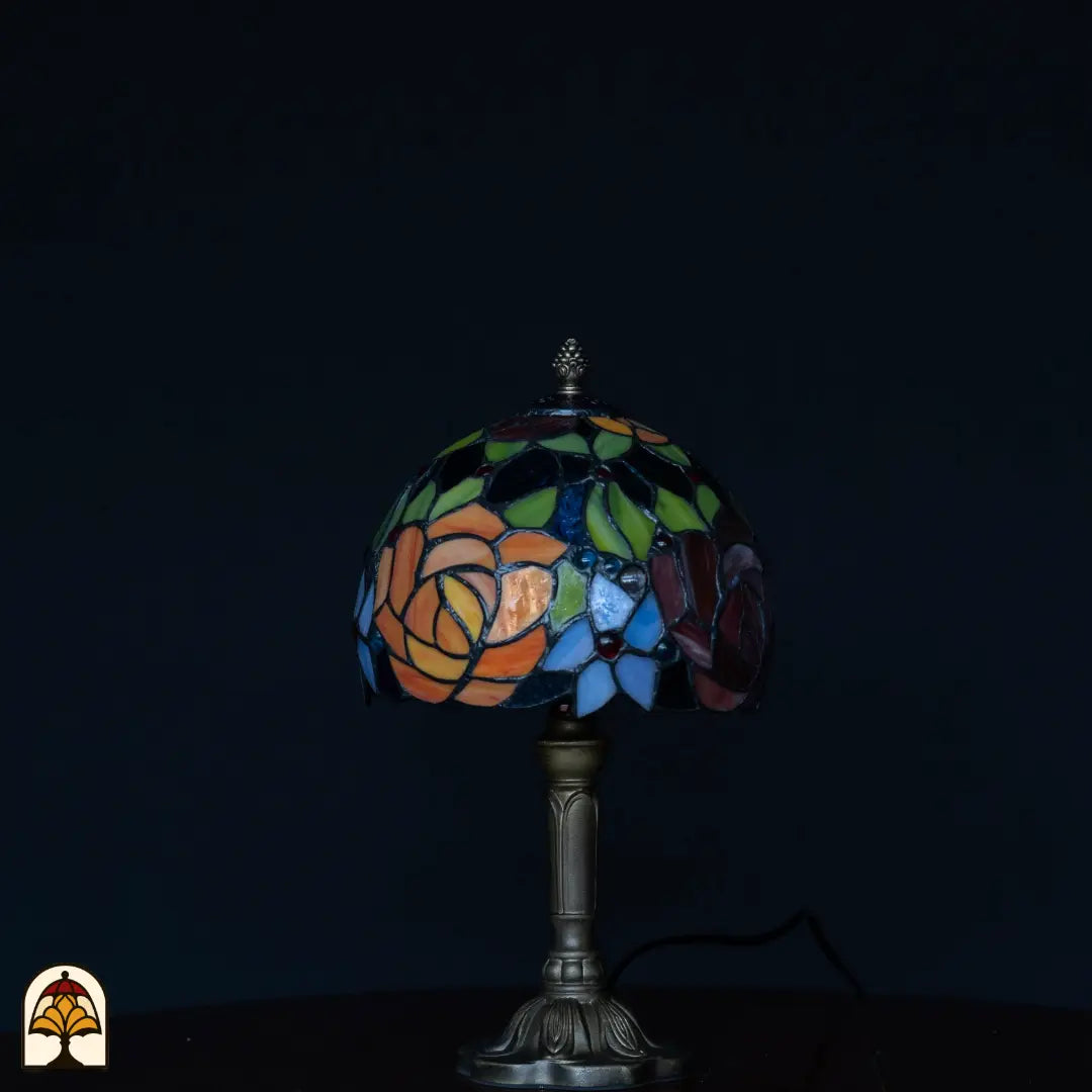 Lampe de table Tiffany Fiore éteinte sur fond sombre — abat-jour avec roses orange, feuilles vertes, fleurs bleues et rose violette, pied colonne en bronze