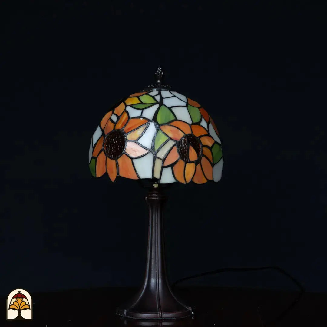 Lampe de table Tiffany Solea éteinte sur fond sombre — abat-jour avec tournesols orange et plages bleues/blanches, feuilles vertes, pied Art Nouveau élancé
