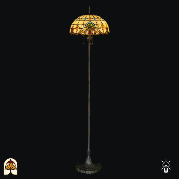 Lampadaire Tiffany Barok allumé sur fond noir — abat-jour dôme baroque crème/ambré avec motifs bleus, verts et jaunes, bord en bronze avec interrupteur à chaînette, long fût avec base large