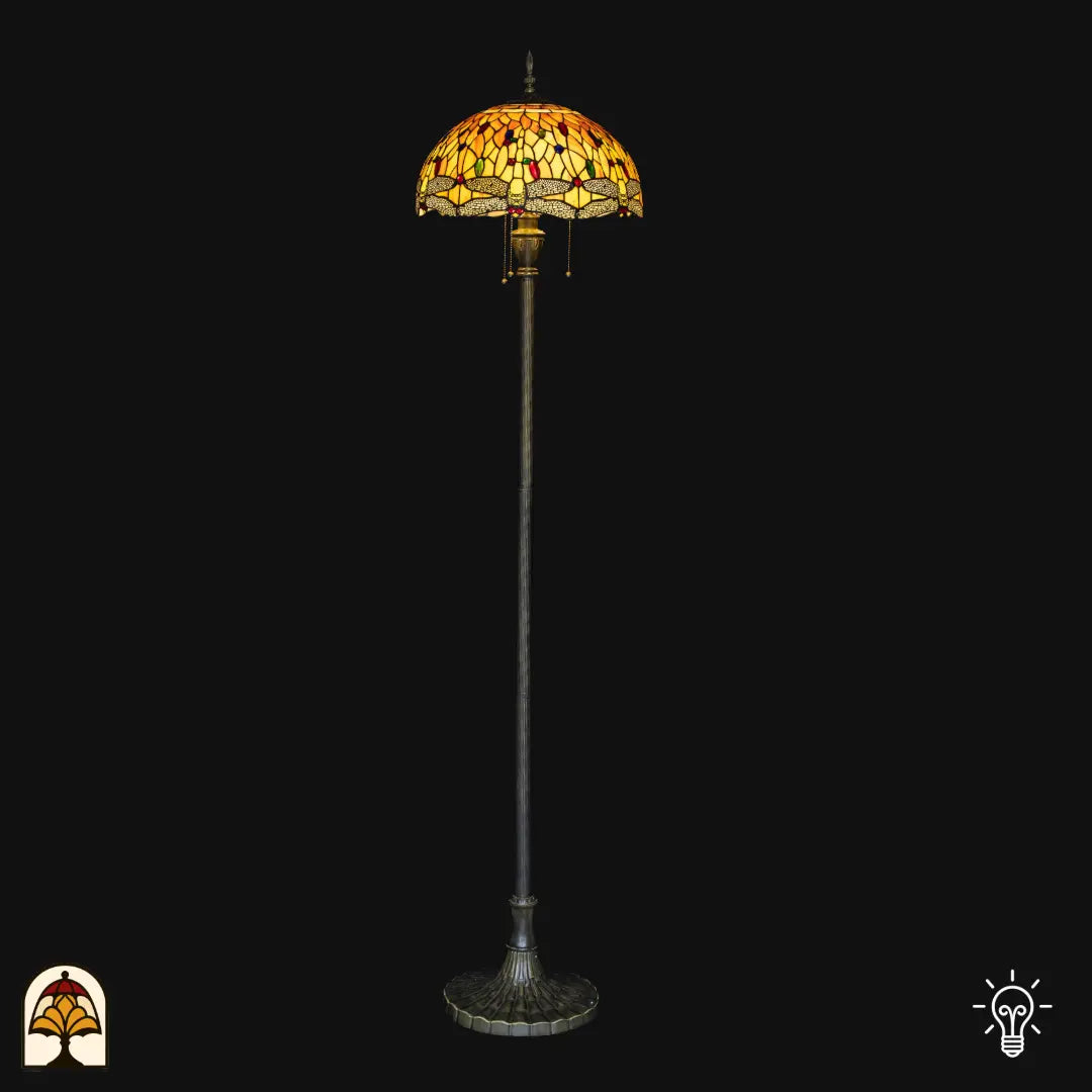 Lampadaire Tiffany Libellula Aurea allumé sur fond noir — abat-jour dôme ambré/jaune avec bordure à libellules, long fût avec base large en bronze, interrupteurs à chaînette