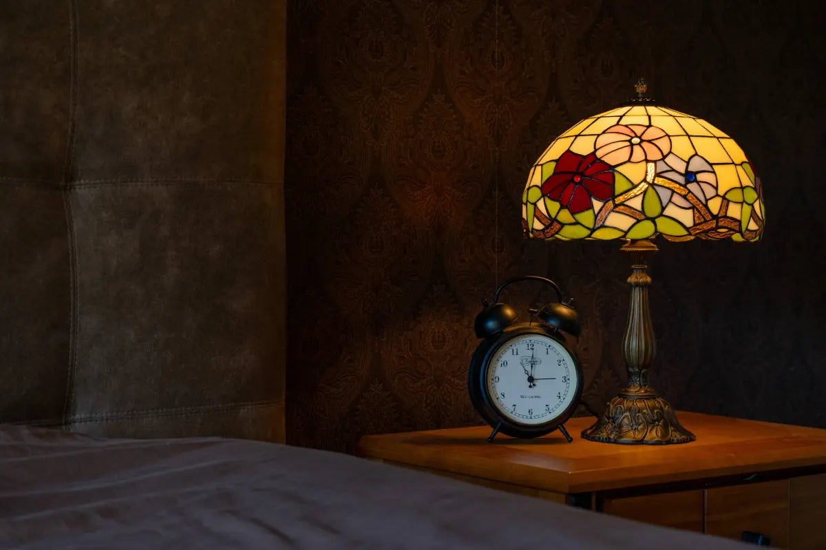 Lampe de table Tiffany à motif floral sur une table de chevet, diffusant une lumière chaude à côté d'un réveil noir classique.