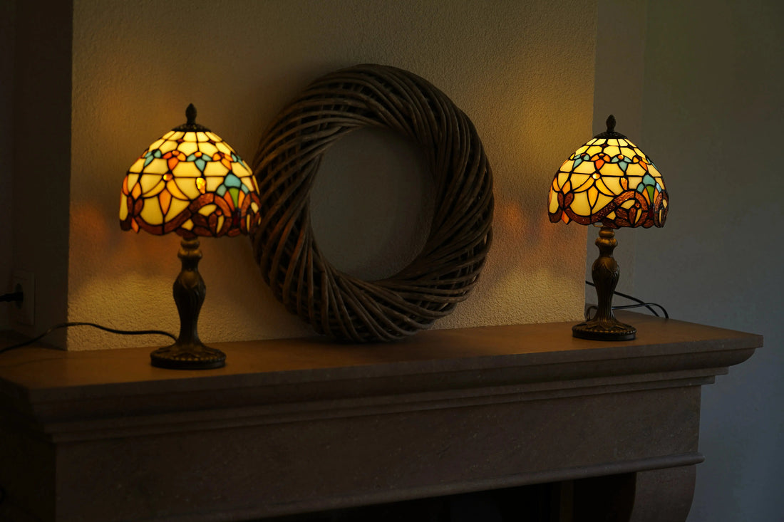 Twee brandende Tiffany tafellampen met warm geel, oranje en turquoise glas staan symmetrisch op een stenen schouw, met een grote ronde rieten wanddecoratie ertussen. De lampen verspreiden zacht, sfeervol licht tegen de wand.