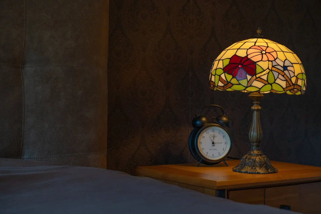 en verlichte Tiffany-stijl tafellamp met een gele kap met groene en bruine bloemmotieven en een bronzen boomstamvoet, geplaatst op een donkerhouten dressoir voor een grote spiegel. Links van de lamp staan rode wijnglazen en een karaf.