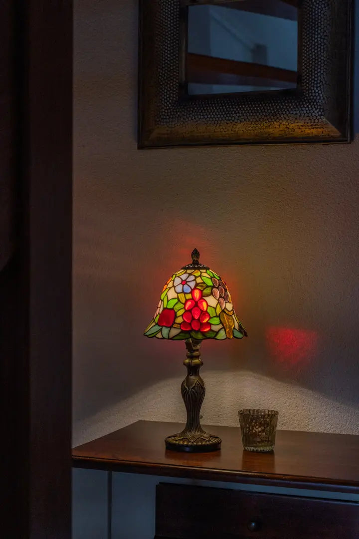 Lampe de table Tiffany colorée en vitrail posée sur un meuble en bois, allumée et projetant une douce lueur d'ambiance sur le mur.