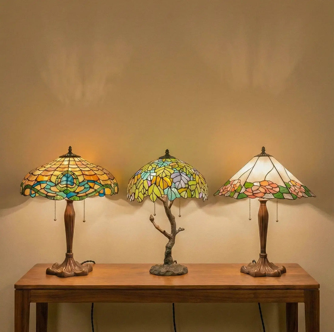 Drie Tiffany tafellampen in verschillende stijlen op een houten tafel, met warm gekleurd glas-in-lood en sfeervol licht tegen een rustige wand.