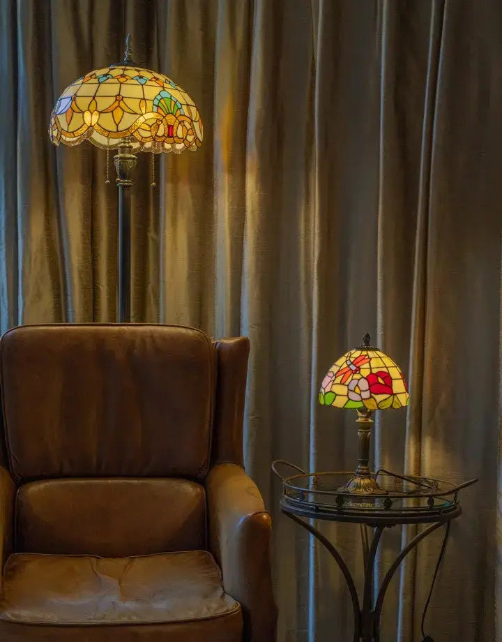 Lampadaire et lampe de table Tiffany dans un coin salon chaleureux, près d'un fauteuil en cuir brun et d'une table basse en verre, abat-jour en vitrail diffusant une lumière douce devant des rideaux beiges.