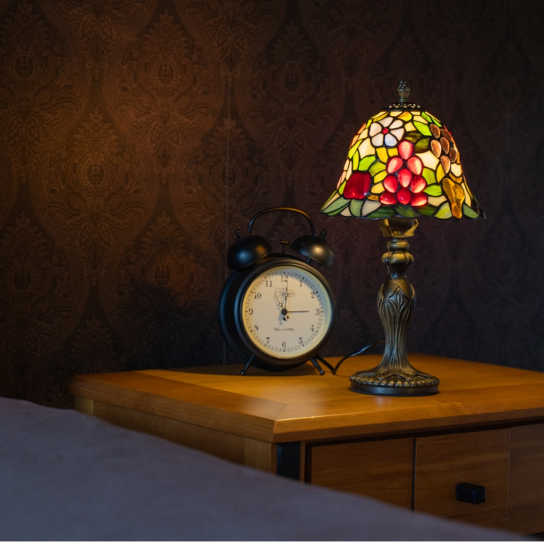 Een Tiffany tafellamp met een bronzen voet en een kleurrijke glas-in-lood kap met fruitmotief in rode, groene en gele tinten, geplaatst op een houten nachtkastje naast een klassieke zwarte wekker.