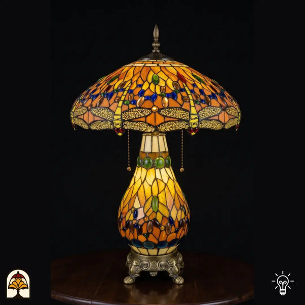 Lampe de table Tiffany allumée, abat-jour en vitrail ambré en dôme avec motifs de libellule le long du bord, cabochons en verre ovales bleus et verts, interrupteur à chaînette ; pied en vitrail en forme de poire et base décorative en bronze à pattes de lion