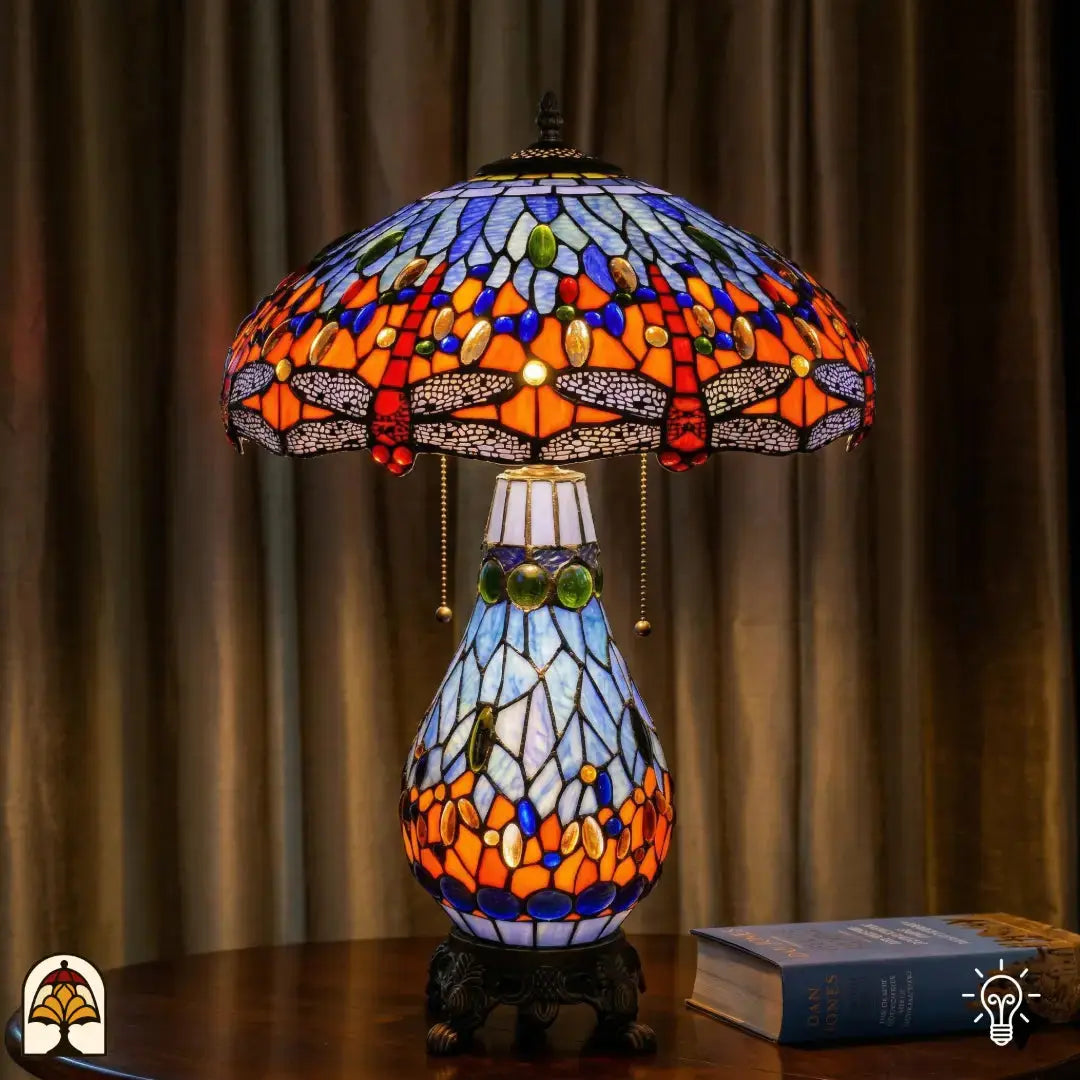 Lampe de table Tiffany allumée sur une table en bois avec rideau en arrière-plan, abat-jour bleu-orange avec motifs de libellule et cabochons en verre bleus, pied en vitrail bleu clair en forme de poire et base en bronze à pattes de lion