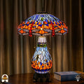 Tiffany lamp tafellamp verlicht op houten tafel met gordijn op de achtergrond, blauw-oranje kap met libellemotieven en blauwe glaskralen, lichtblauwe glas-in-lood peervormige voet en bronzen leeuwenpootvoet