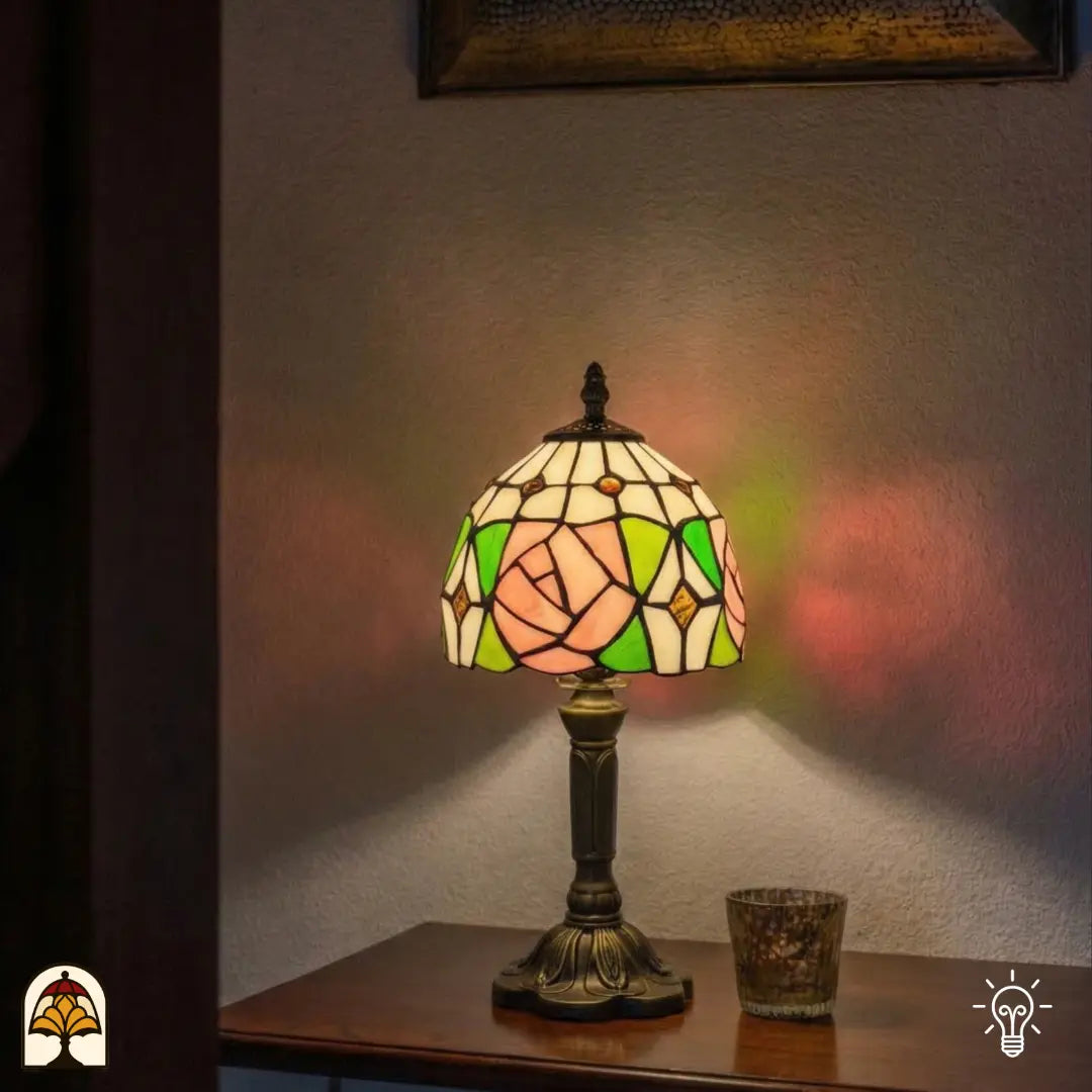 Lampe de table Tiffany Rosalia Verde allumée sur un buffet en bois à côté d'une bougie d'ambiance, rose rose et feuilles vertes illuminées, reflets de lumière colorés sur le mur
