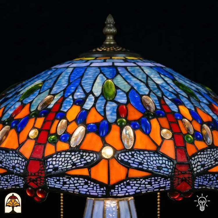 Detailopname van tiffany lamp kapbovenzone verlicht, blauw en lichtblauw glas met blauwe, groene en amber ovale glaskralen in een band, puntfinial bovenaan zichtbaar