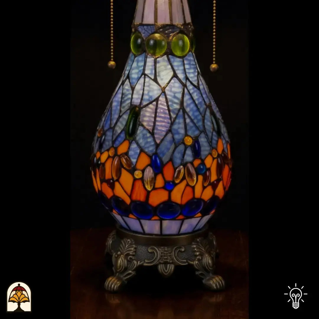 Gros plan de lampe Tiffany, pied piriforme en vitrail allumé, verre bleu clair avec cabochons en verre ovales verts, bleus et orange, chaîne de l'interrupteur visible et pied décoratif en bronze à pattes de lion
