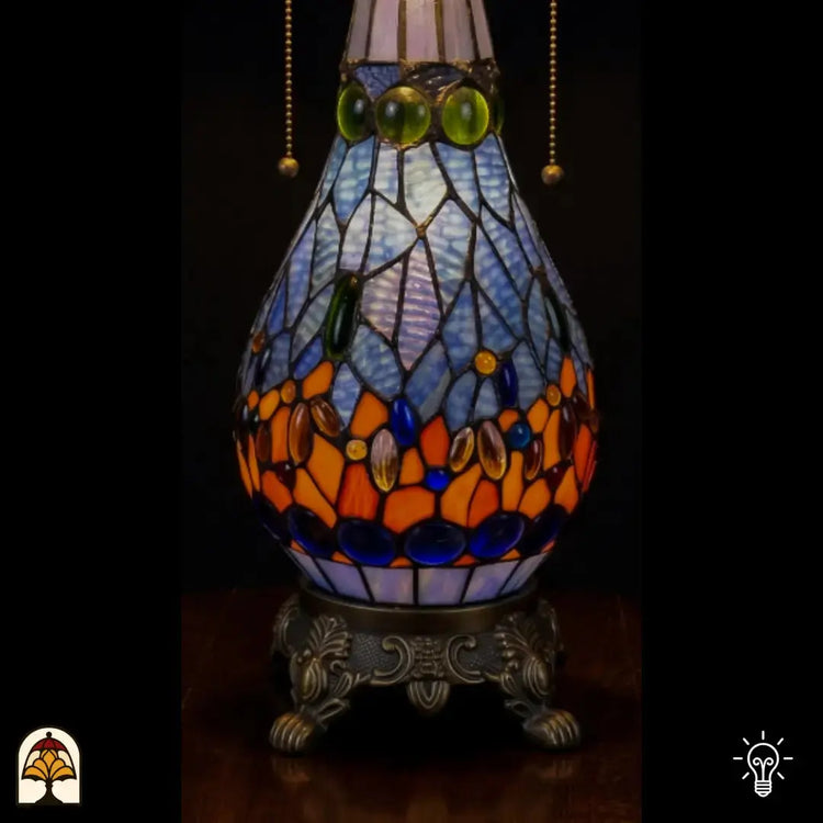 Detailopname van tiffany lamp glas-in-lood peervormige voet verlicht, lichtblauw glas met groene, blauwe en oranje glaskralen, trekketting zichtbaar en bronzen siervoet met leeuwenpoten