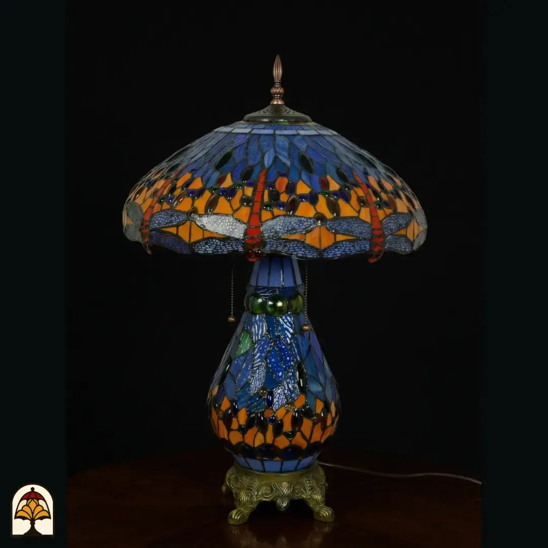 Lampe de table Tiffany éteinte, abat-jour en dôme avec verre bleu et motifs de libellule le long du bord, cabochons en verre orange et rouges, pied en vitrail bleu en forme de poire et base en bronze à pattes de lion