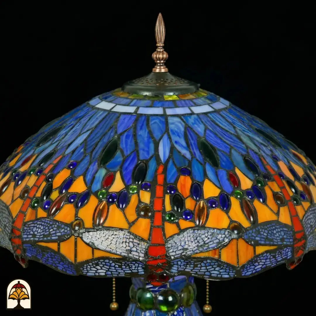 Gros plan de lampe Tiffany, zone supérieure de l'abat-jour éteinte, verre bleu et bleu clair avec cabochons en verre ovales bleus, verts et ambrés en bandes, fleuron pointu et plaque d'aération en bronze visibles en haut