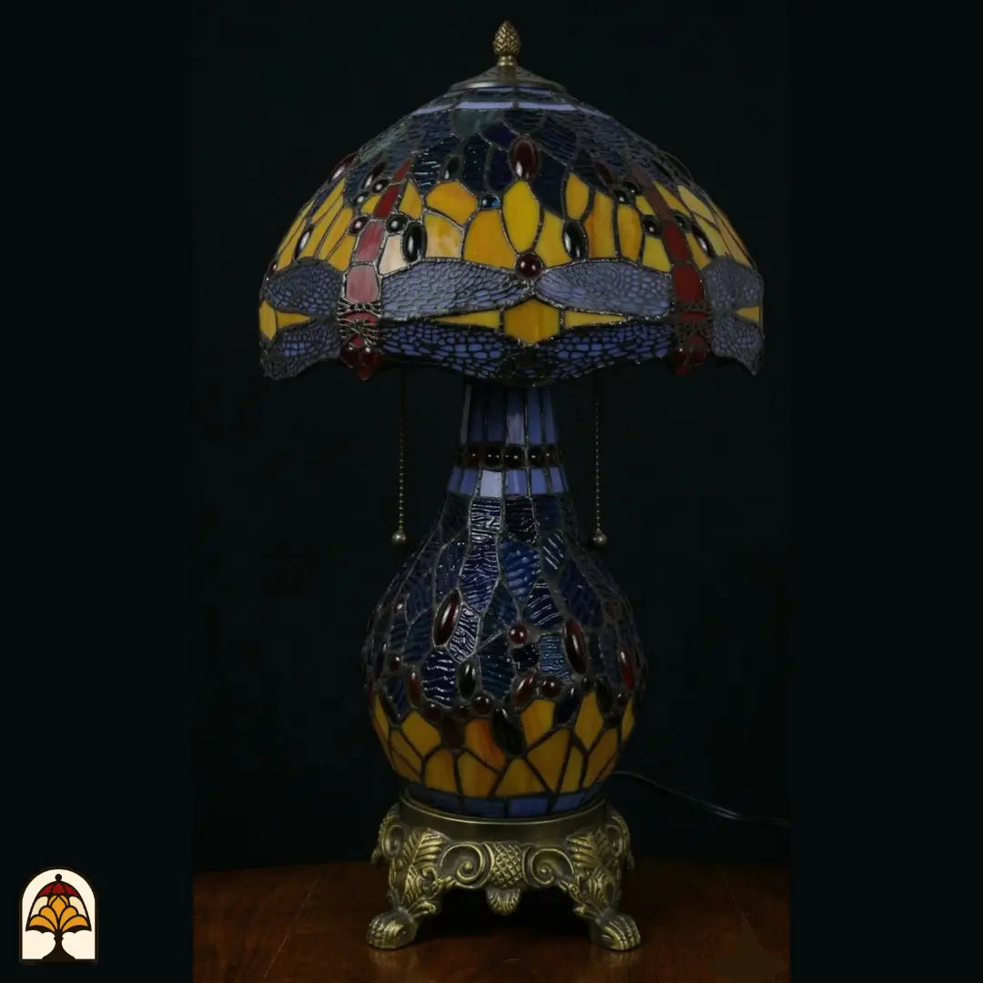 Lampe de table Tiffany Libellula Azurea ⌀30 cm éteinte sur fond sombre – abat-jour dôme à motif de libellule en vitrail bleu et ambré, pied boule en verre avec interrupteurs à chaînette