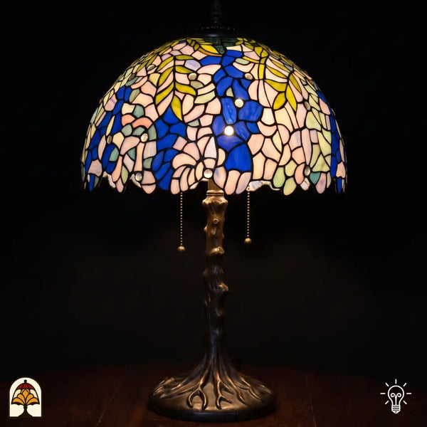 Lampe de table Tiffany Wisteria Celeste ⌀40 cm allumée sur fond noir – abat-jour en vitrail glycine bleu et crème, pied en forme de tronc d'arbre en bronze avec racines
