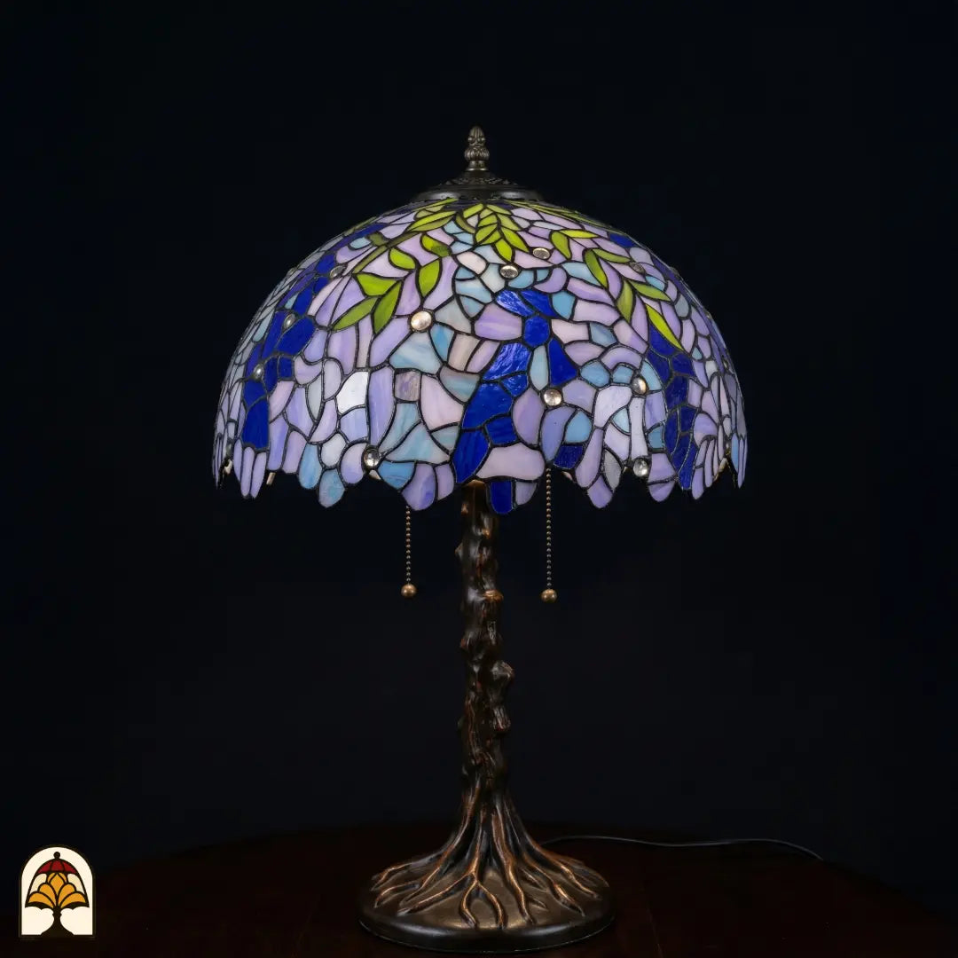 Lampe de table Tiffany Wisteria Celeste éteinte sur fond sombre avec fleurs de glycine lilas-bleu et blanches, feuilles vertes et pied en forme de tronc d'arbre en bronze avec racines