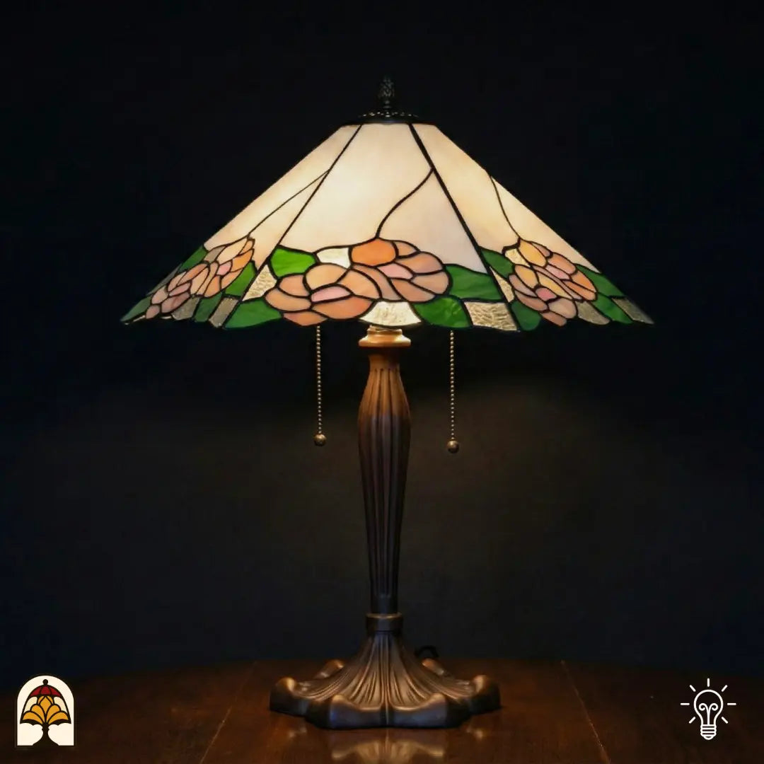 Lampe de table Tiffany Alba Sereno allumée avec large abat-jour trapézoïdal, partie supérieure blanche et crème avec roses roses et feuilles vertes le long du bord, pied en bronze cannelé avec interrupteurs à chaînette, fond sombre