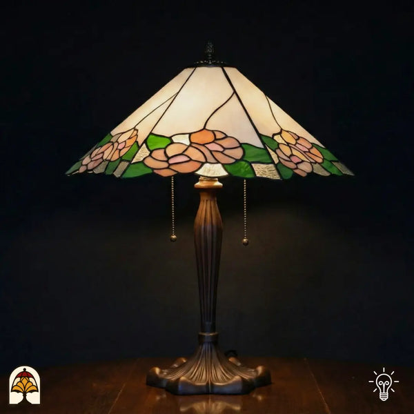 Lampe de table Tiffany Alba Sereno allumée avec large abat-jour trapézoïdal, partie supérieure blanche et crème avec roses roses et feuilles vertes le long du bord, pied en bronze cannelé avec interrupteurs à chaînette, fond sombre