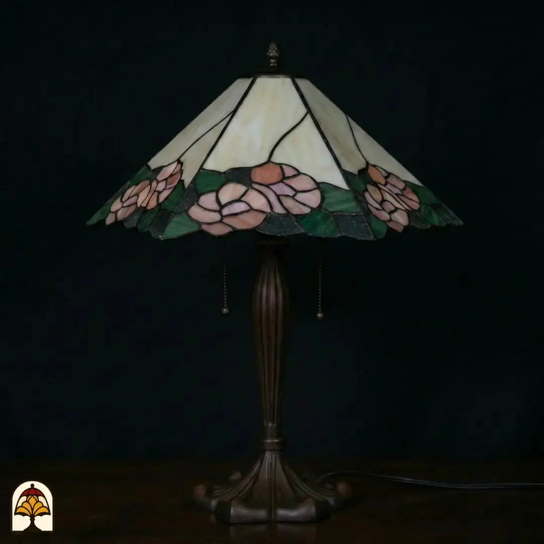 Lampe de table Tiffany Alba Sereno éteinte sur fond bleu foncé, large abat-jour trapézoïdal avec roses roses et feuilles vertes, pied en bronze avec interrupteurs à chaînette