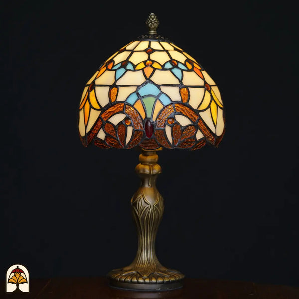 Lampe de table Tiffany Baroque ⌀20 cm allumée sur fond noir – abat-jour en dôme crème avec feuillages baroques ambrés, formes arquées bleu clair et pierre précieuse rouge, pied Art nouveau en bronze ouvragé